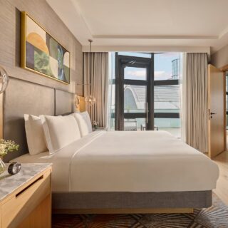 Modern hotel room with a king bed, soft white linens, and a wood-paneled headboard under round glass pendant lights. A balcony door with sheer curtains lets in natural light.