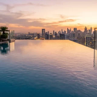 Rooftop infinity pool at sunset with lounge chairs and potted plants, overlooking a city skyline.