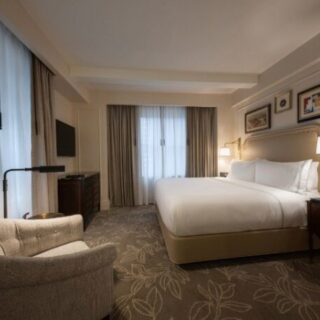 Spacious hotel room with a large bed, two nightstands, lamps, and an upholstered chair near a windowed wall.