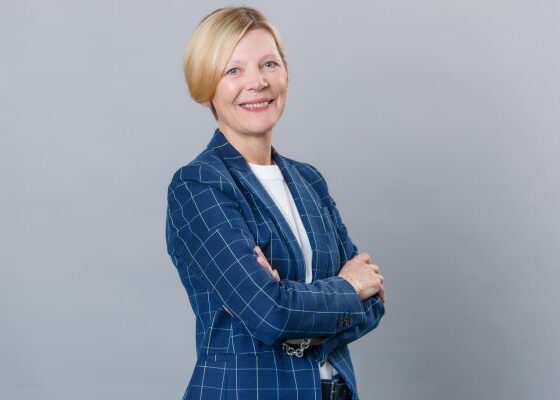 Professional woman with short blonde hair in a blue checkered blazer, arms crossed, smiling at the camera.