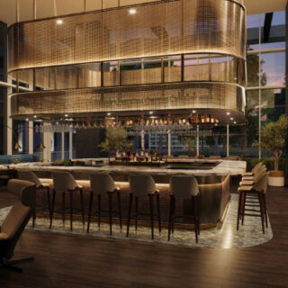 Interior of a contemporary hotel bar with a curved marble counter, beige bar stools, and a large metal mesh circular chandelier overhead, near floor-to-ceiling windows at dusk.