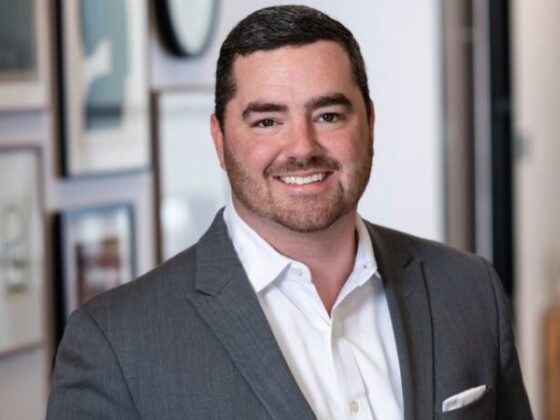 Smiling man in a gray suit and white shirt standing in an office hallway/office setting.