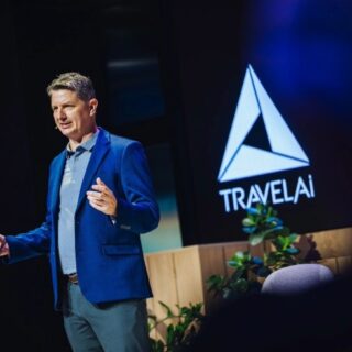 Male presenter in a blue blazer speaks on stage with a TravelAI logo on the screen behind him.