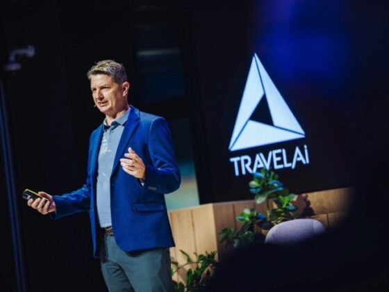 Male presenter in a blue blazer speaks on stage with a TravelAI logo on the screen behind him.