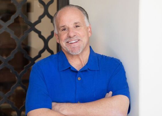 Smiling man with gray hair in a blue polo shirt stands with arms crossed beside a doorway behind a decorative screen.
