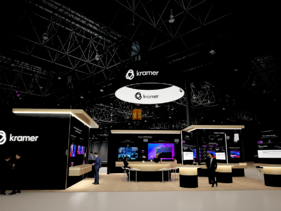 Trade show booth with Kramer branding, large black walls, circular suspended sign, and multiple product displays with people talking around a curved counter.
