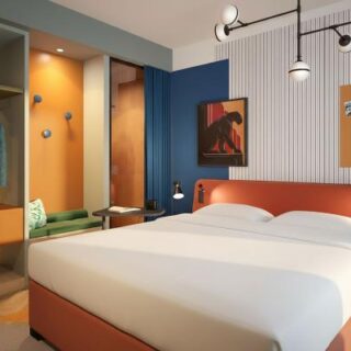 Modern hotel room with a large orange headboard, white bed, and seating nook by an orange wall alcove