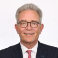 Portrait of a smiling older man with gray hair and glasses, wearing a navy pinstripe suit and red tie against a white background, in a professional headshot.
