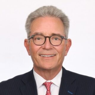 Portrait of a smiling older man with gray hair and glasses, wearing a navy pinstripe suit and red tie against a white background, in a professional headshot.