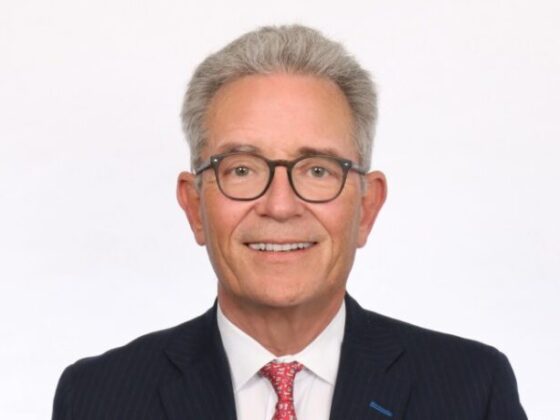 Portrait of a smiling older man with gray hair and glasses, wearing a navy pinstripe suit and red tie against a white background, in a professional headshot.