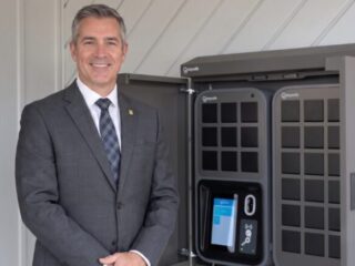 Businessman in a gray suit stands beside a large gray vehicle charging kiosk, smiling at the camera.