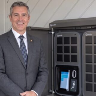 Businessman in a gray suit stands beside a large gray vehicle charging kiosk, smiling at the camera.