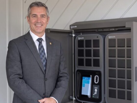 Businessman in a gray suit stands beside a large gray vehicle charging kiosk, smiling at the camera.