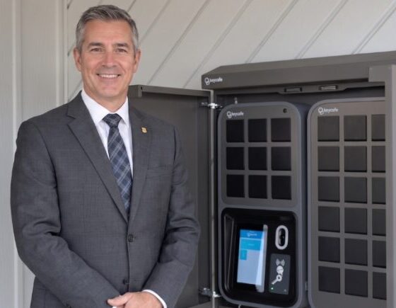 Businessman in a gray suit stands beside a large gray vehicle charging kiosk, smiling at the camera.