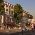 Grand white building with arched windows along a tree-lined street at dusk, a red double-decker bus parked near the curb, and a blue car ahead; storefront signs visible including 'Citizen'.