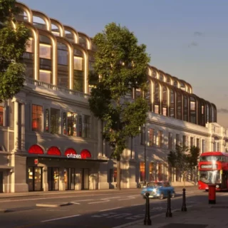 Grand white building with arched windows along a tree-lined street at dusk, a red double-decker bus parked near the curb, and a blue car ahead; storefront signs visible including 'Citizen'.