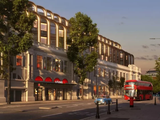 Grand white building with arched windows along a tree-lined street at dusk, a red double-decker bus parked near the curb, and a blue car ahead; storefront signs visible including 'Citizen'.