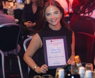 Smiling woman at a crowded awards event holding a framed certificate that reads 'One to Watch' at a tableside banquet.