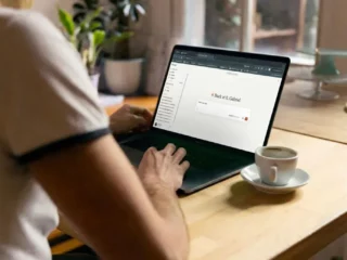 Person typing on a laptop at a wooden table with a cup of coffee nearby; the laptop screen shows a login page with the message 'Back at it, Gabriel'.