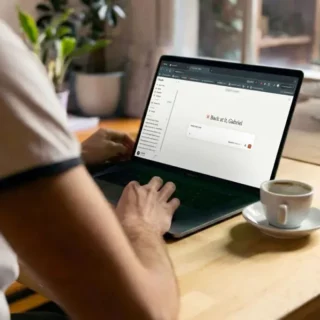 Person typing on a laptop at a wooden table with a cup of coffee nearby; the laptop screen shows a login page with the message 'Back at it, Gabriel'.