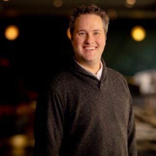 Smiling man in a dark speckled sweater posing in a warmly lit indoor space.