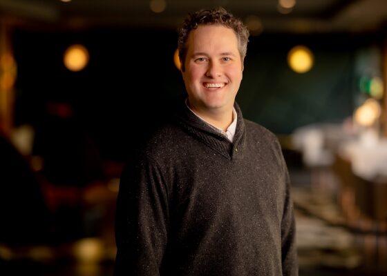 Smiling man in a dark speckled sweater posing in a warmly lit indoor space.