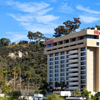 Sheraton hotel building on hillside with palm trees in the foreground under a clear blue sky.