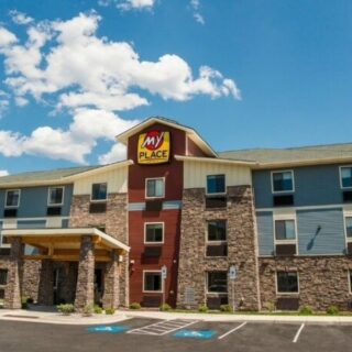 Exterior view of a My Place hotel with stone columns, blue siding, and a covered entry on a sunny day