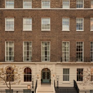 Brown brick townhouse facade with white-framed sash windows and a white entryway, blue front door, and stairs flanked by black iron railings.
