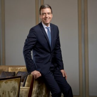 Man in a navy suit and blue tie stands beside a beige sofa in a modern office lobby, smiling at the camera.