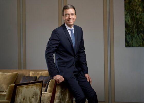 Man in a navy suit and blue tie stands beside a beige sofa in a modern office lobby, smiling at the camera.