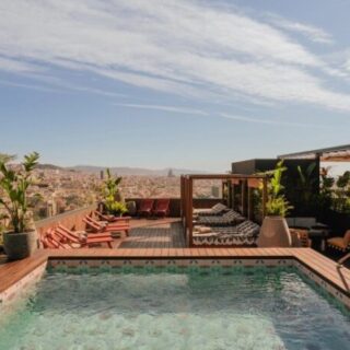 Rooftop pool on a wooden deck with lounge chairs, planters, and a shaded seating area overlooking a city skyline.