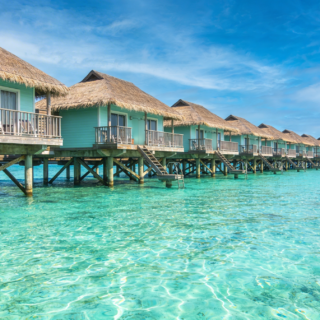 Overwater bungalows with thatched roofs on stilts line a turquoise lagoon under a bright blue sky.