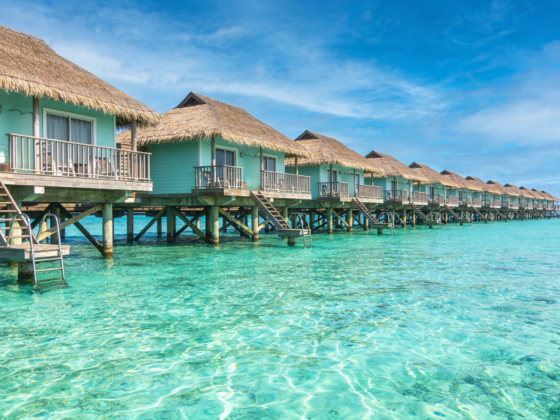 Overwater bungalows with thatched roofs on stilts line a turquoise lagoon under a bright blue sky.
