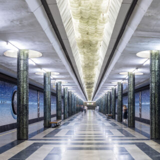 Symmetrical underground metro platform with blue shutter doors, glossy green marble columns, and a checkered floor pattern nearby.