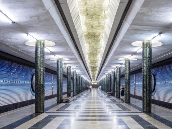 Symmetrical underground metro platform with blue shutter doors, glossy green marble columns, and a checkered floor pattern nearby.