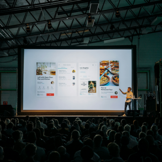Speaker on stage presenting a projector screen with multiple app mockups to a large audience under dim lighting in a conference hall.