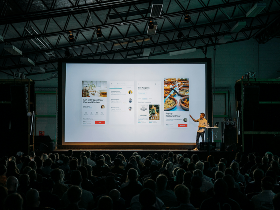 Speaker on stage presenting a projector screen with multiple app mockups to a large audience under dim lighting in a conference hall.