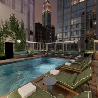Rooftop pool area with green cushioned lounge chairs, towels, and small tables, overlooking a city skyline at dusk.