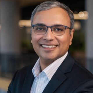 Professional headshot of a smiling man in a dark blazer and light shirt, wearing glasses, in a modern office setting