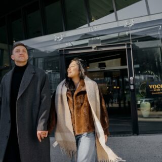 Couple holding hands and walking out of a glass-fronted building entrance in stylish coats.