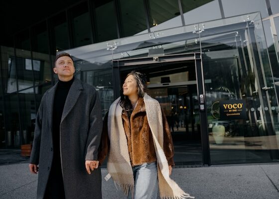 Couple holding hands and walking out of a glass-fronted building entrance in stylish coats.