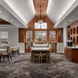 Bright communal dining area with a central high table, chairs, and a hanging glass pendant light near wood cabinets.
