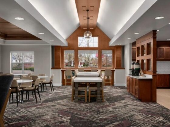 Bright communal dining area with a central high table, chairs, and a hanging glass pendant light near wood cabinets.