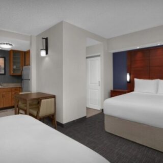 Hotel suite with two beds, a dark wood headboard wall, and a compact kitchenette nearby.