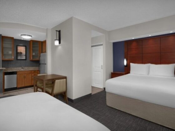 Hotel suite with two beds, a dark wood headboard wall, and a compact kitchenette nearby.