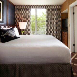 Hotel room featuring a large white bed with a dark headboard, patterned curtains, a bedside lamp, window view, dresser with a TV, and an open door.
