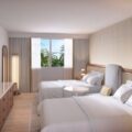 Hotel room with two neatly made white beds, a bedside table, and a large window showing palm trees outside.