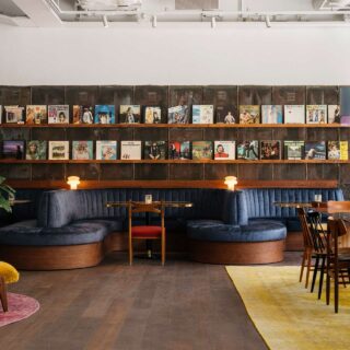 Cafe interior with dark wood and blue upholstered seating, vinyl records on a wall display, plants, and teal pendant lamps by large windows.