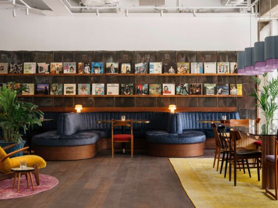 Cafe interior with dark wood and blue upholstered seating, vinyl records on a wall display, plants, and teal pendant lamps by large windows.